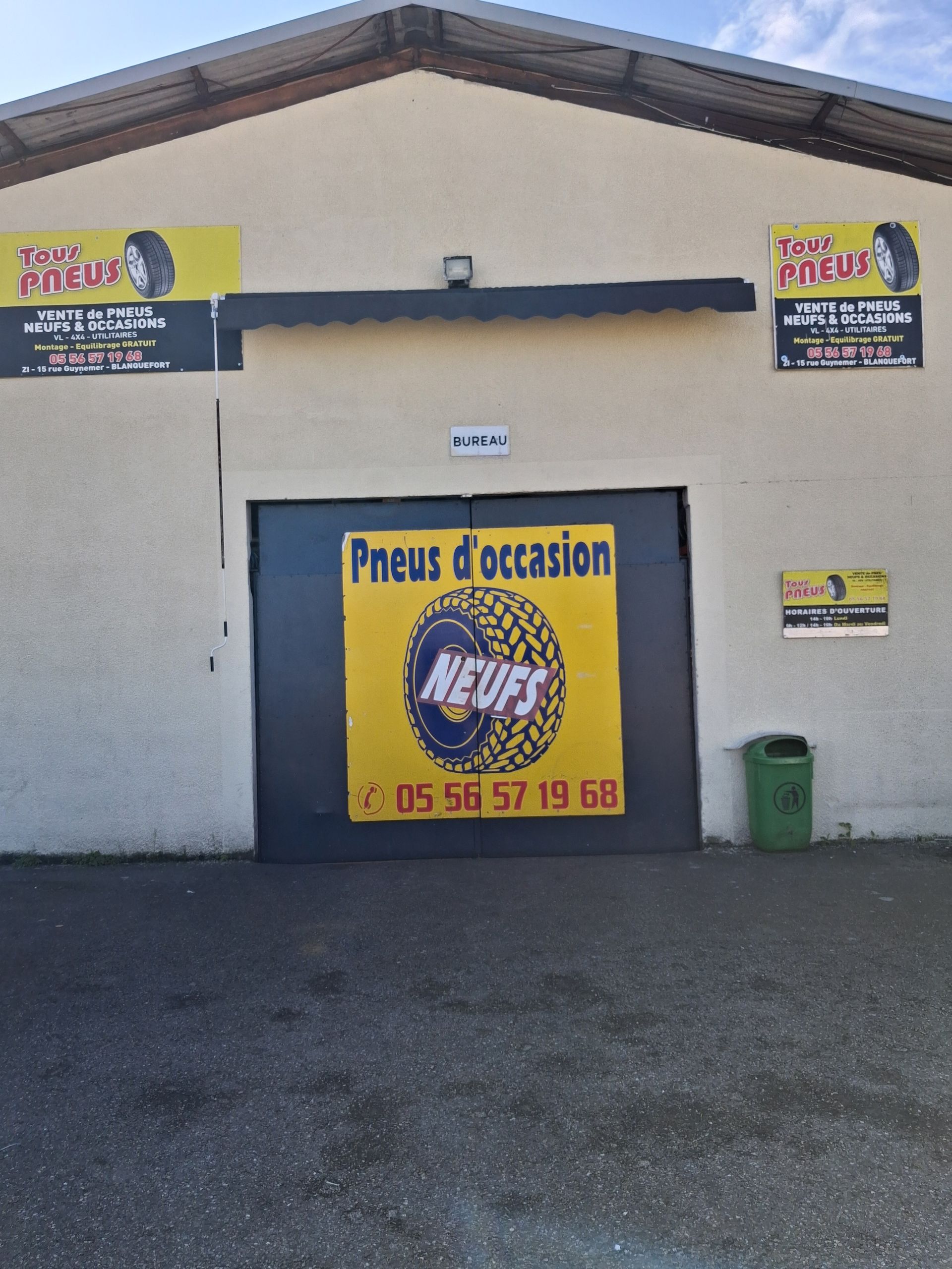 L'extérieur d'un magasin de pneus avec une enseigne jaune et un numéro de téléphone, et de grandes portes d'entrepôt en métal sombre.