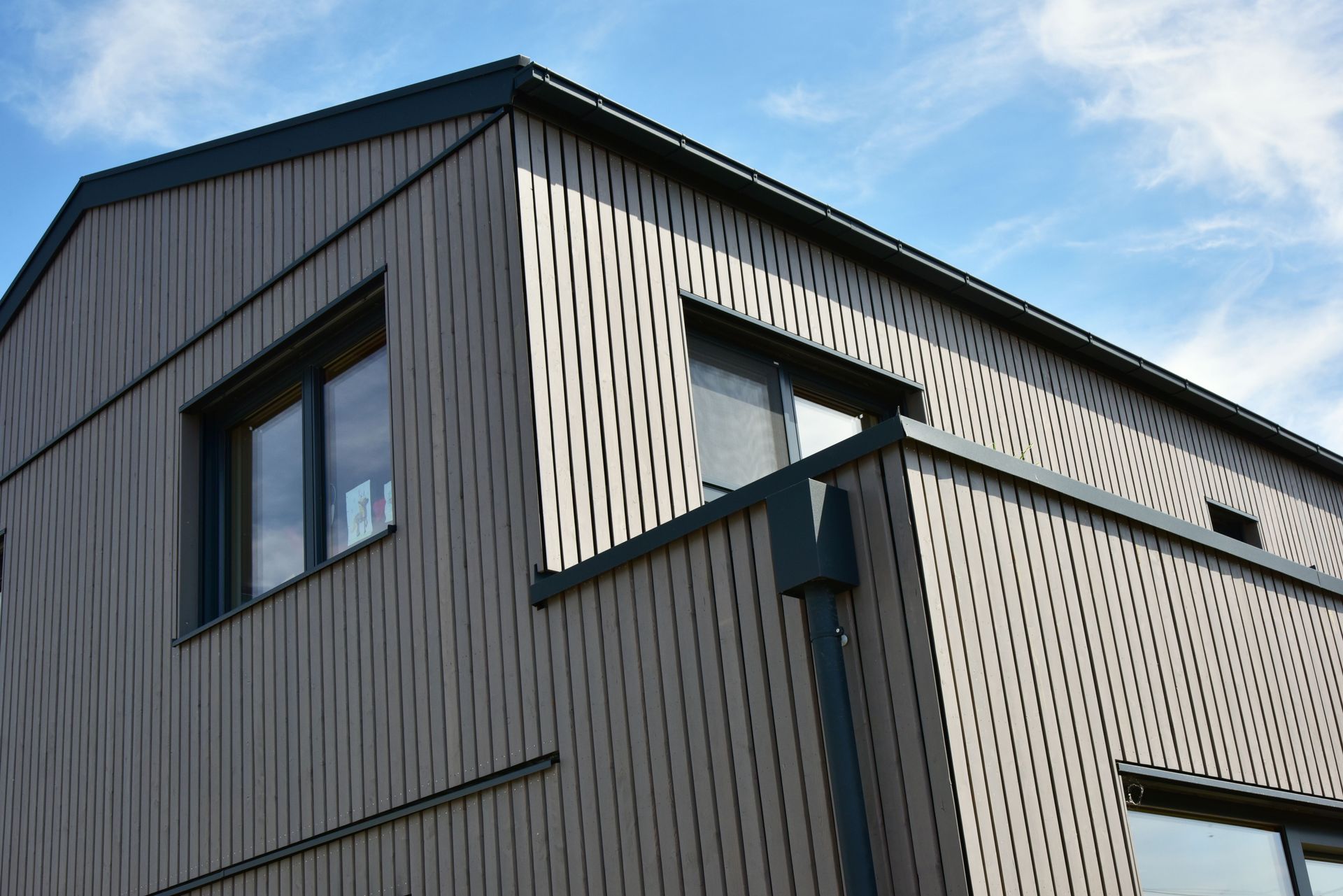 Façade d'un bâtiment moderne avec bardage gris, boiseries sombres et fenêtres se détachant sur un ciel bleu.