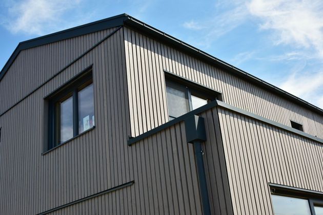 Façade d'un bâtiment moderne avec bardage gris, boiseries sombres et fenêtres se détachant sur un ciel bleu.