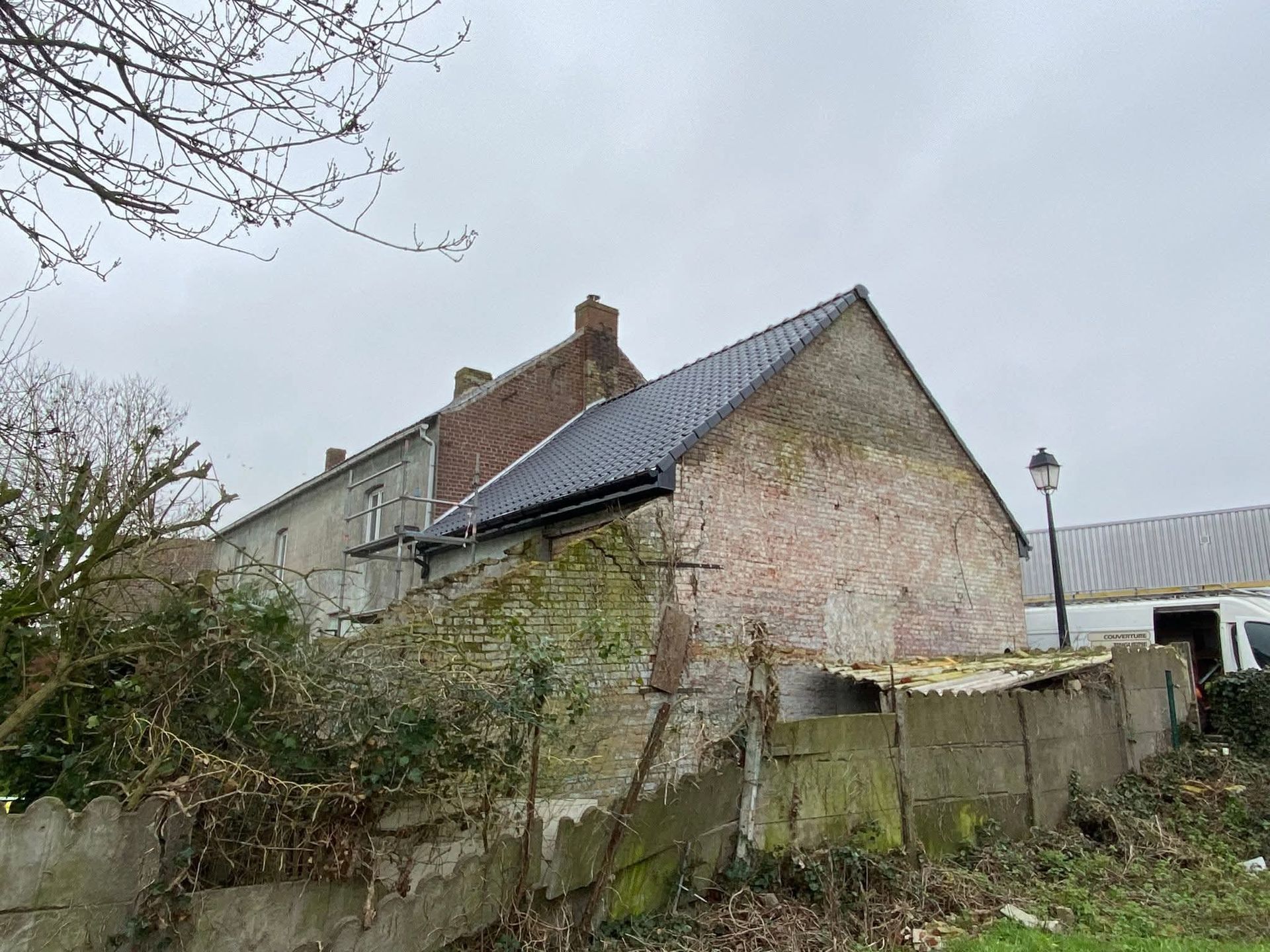Vieux bâtiment en briques, au toit sombre et à la clôture en béton patiné, sous un ciel couvert.