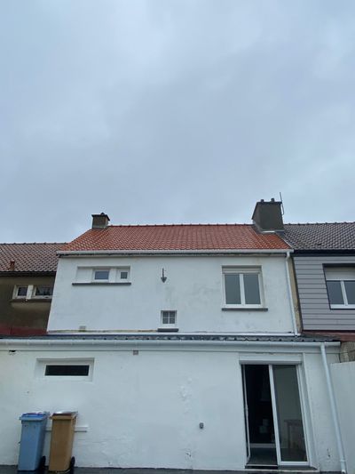 Maison blanche au toit de tuiles rouges sur fond de ciel nuageux. Une poubelle bleu et beige se trouve devant.