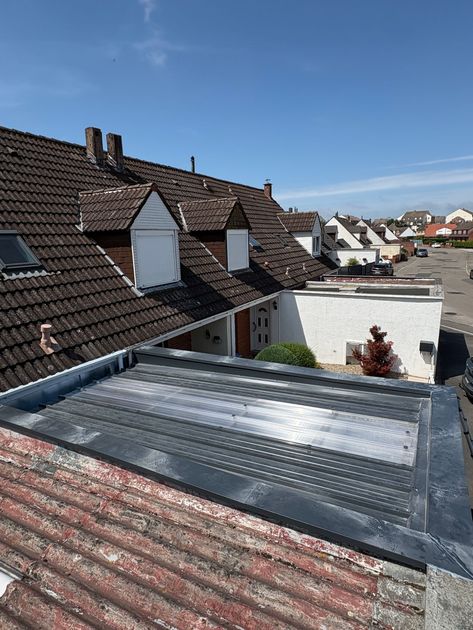 Lucarne sur un toit de tuiles. Bardage gris, encadrements rouges et fenêtre à cadre blanc sur fond de ciel nuageux.