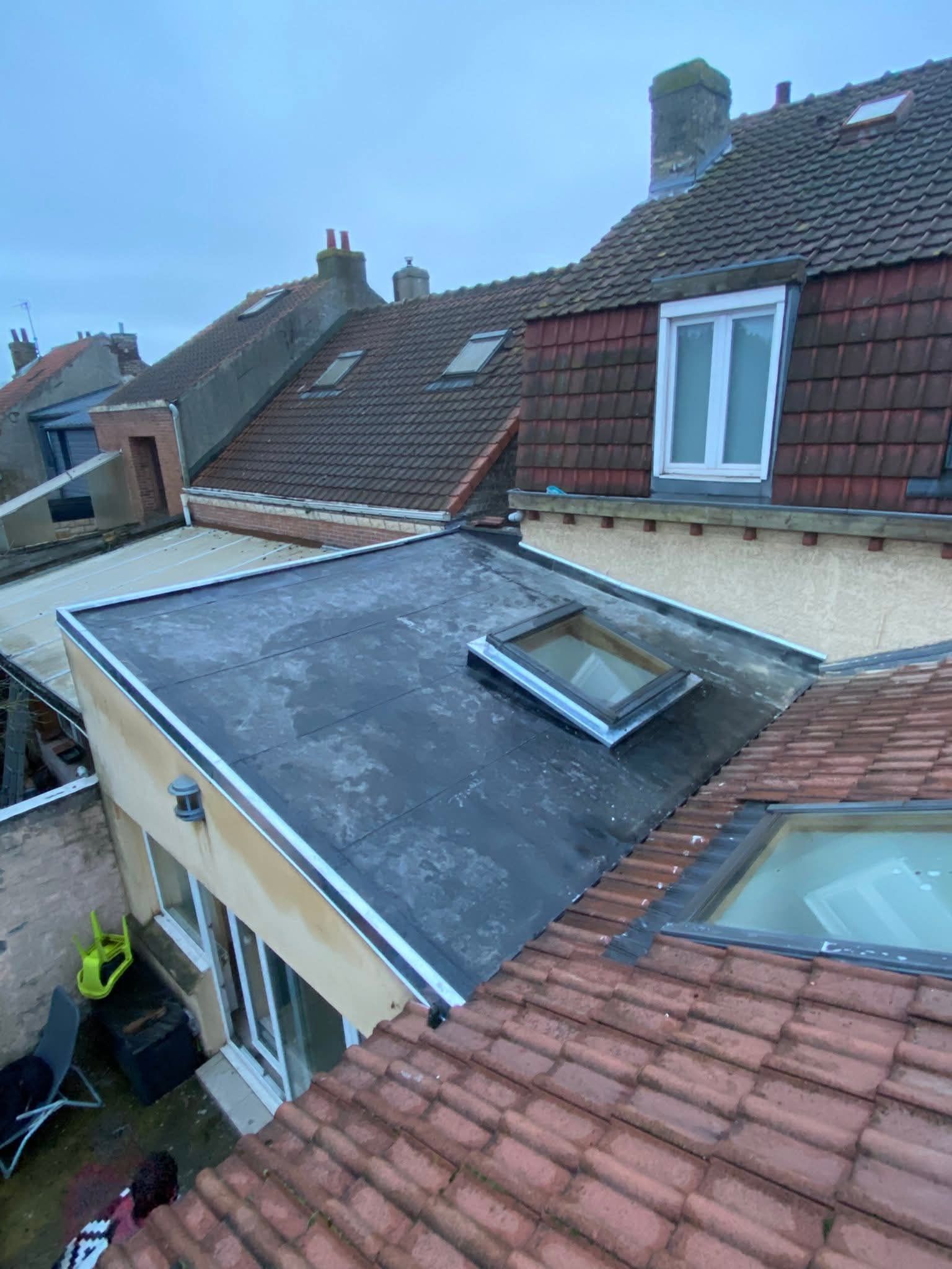 Toits de bâtiments ; toit plat en asphalte foncé avec puits de lumière ; toits de tuiles rouges en arrière-plan.