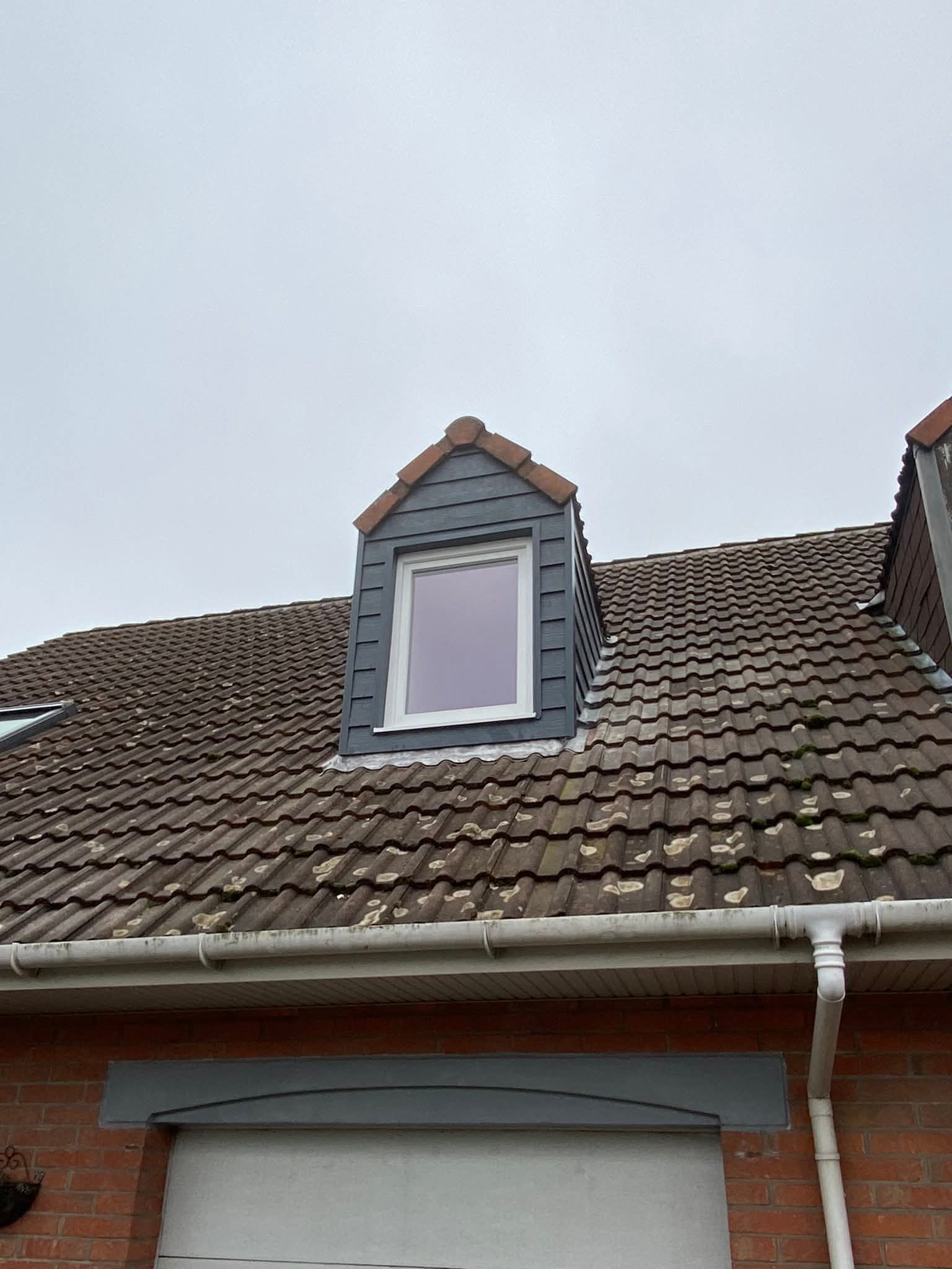 Lucarne sur un toit de tuiles. Bardage gris, encadrements rouges et fenêtre à cadre blanc sur fond de ciel nuageux.