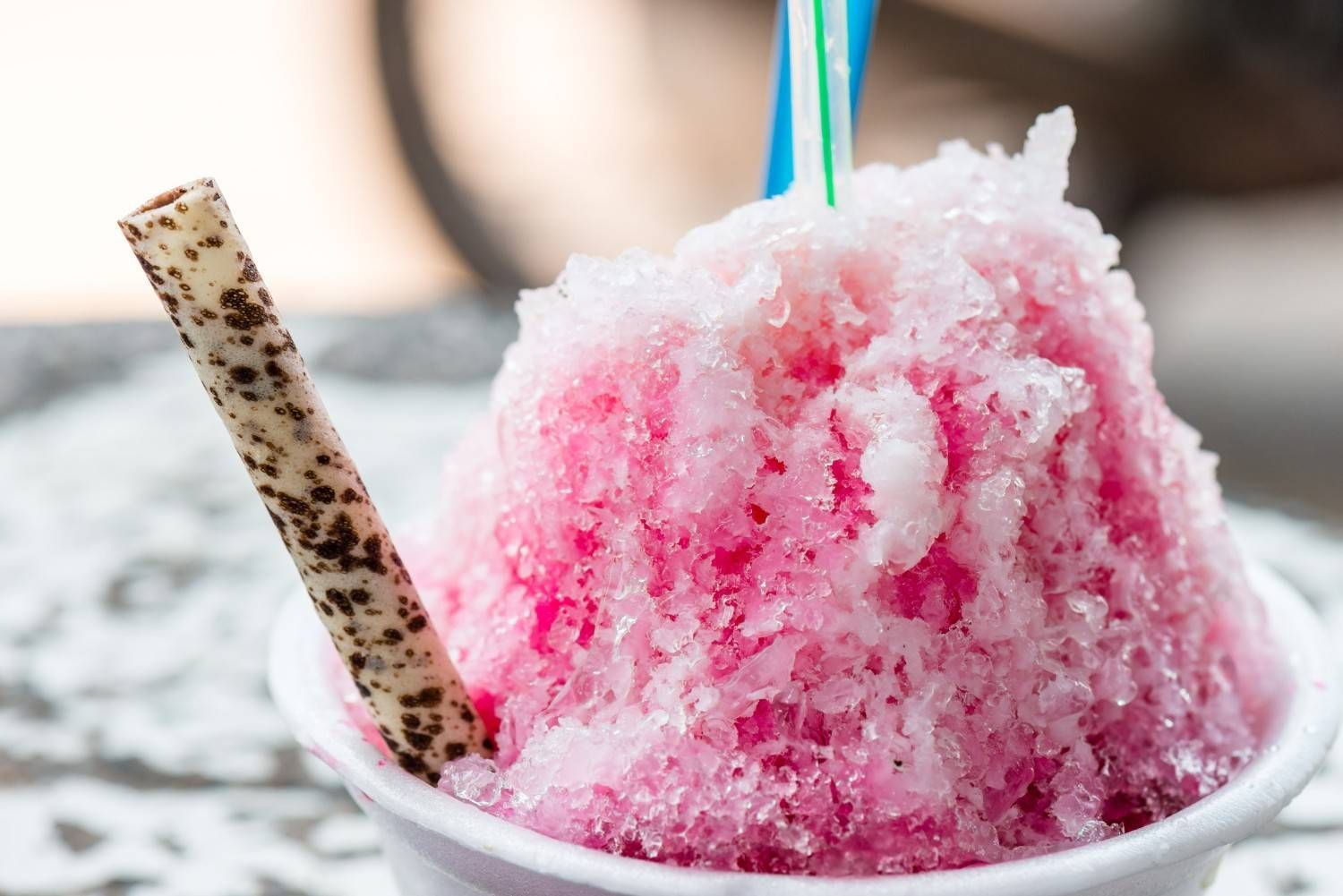 Hielo raspado rosado en vaso blanco, con palito de oblea y dos pajitas.
