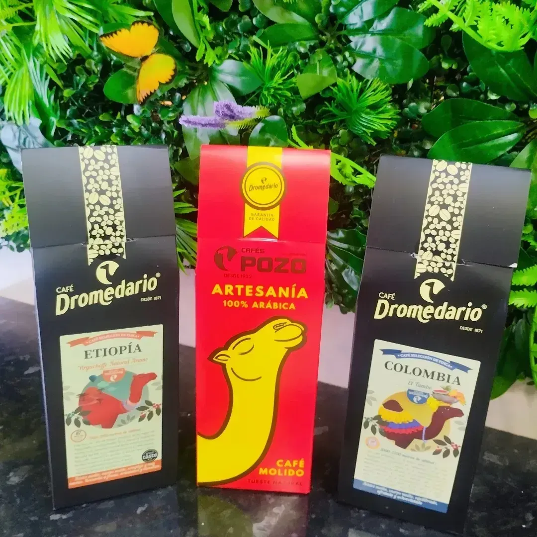 Tres paquetes de café: uno rojo y dos negros con ilustraciones coloridas, sobre un fondo de plantas verdes.