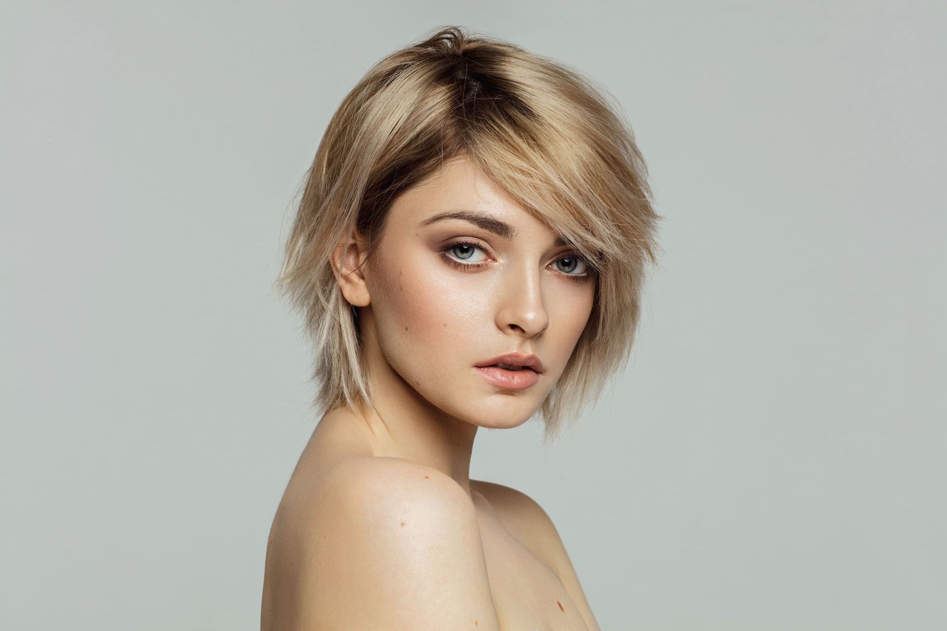 Mujer rubia con cabello corto en capas que mira por encima del hombro con una expresión neutra sobre un fondo gris.