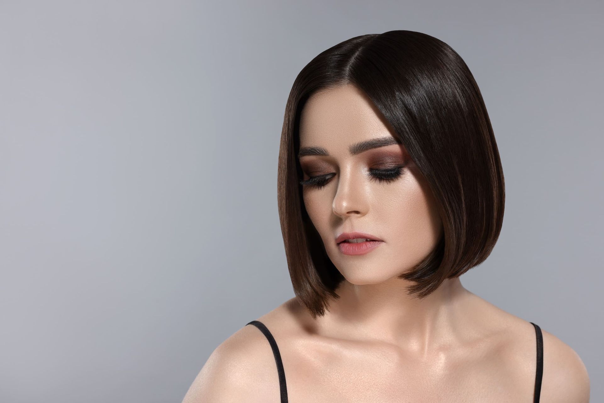 Mujer con peinado bob liso y oscuro y maquillaje de ojos ahumados mirando hacia abajo sobre un fondo gris.