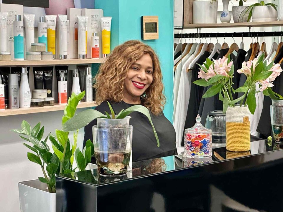 Mujer de cabello rizado sonríe tras un mostrador negro. Al fondo productos en los estantes. Plantas y flores decoran.