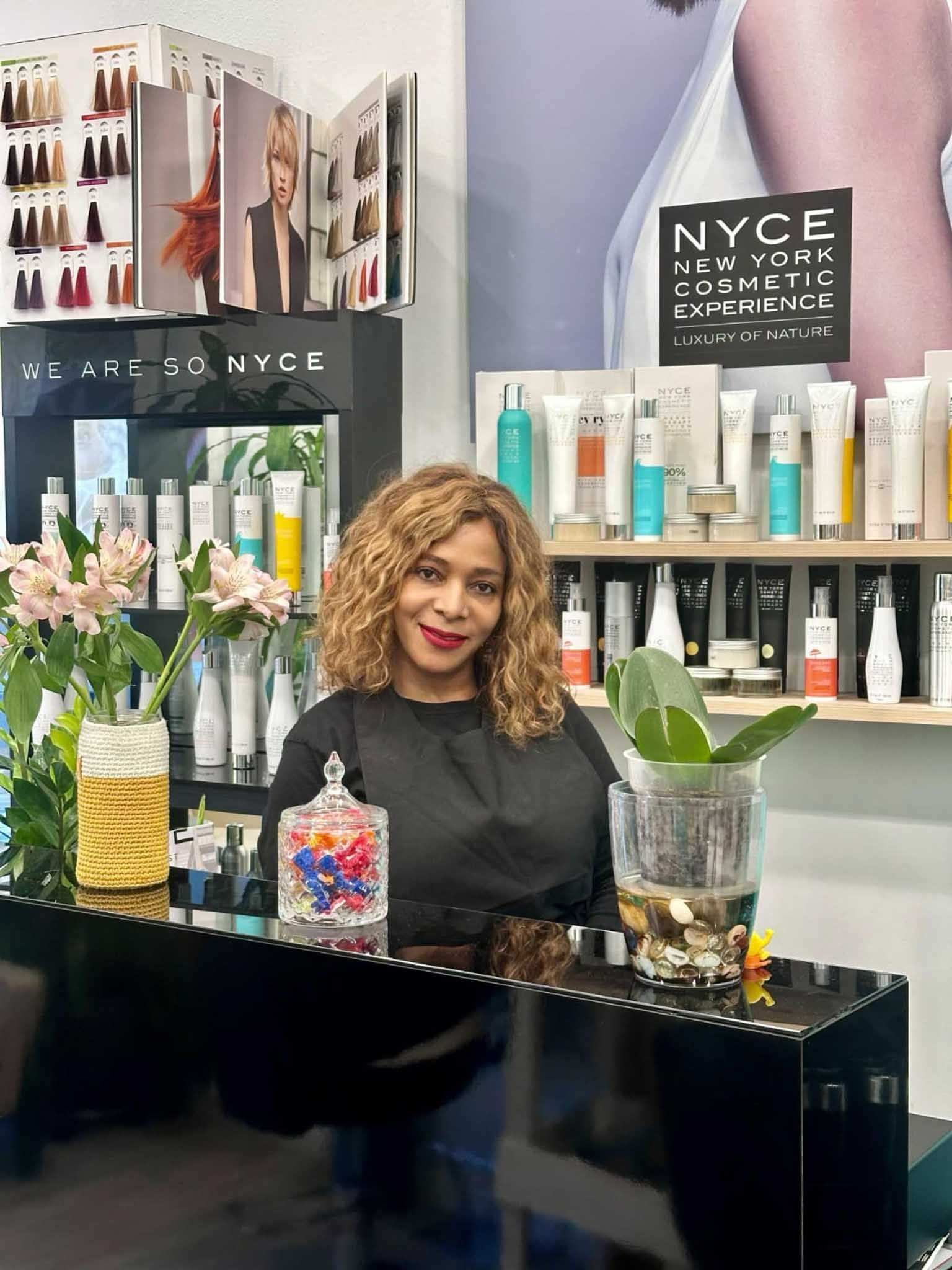 Mujer de cabello rizado detrás de un mostrador de peluquería lleno de productos y  Sonríe en un espacio luminoso.