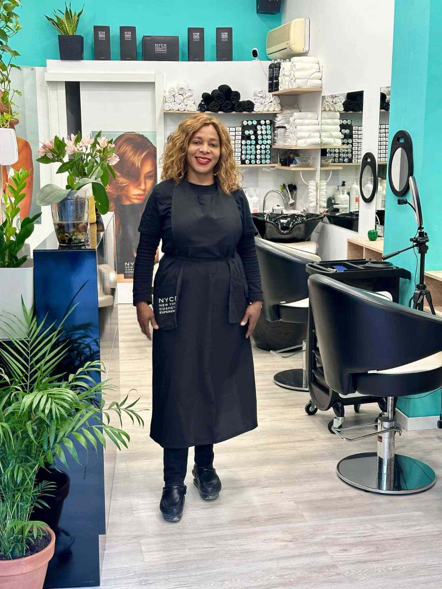 Una mujer con delantal negro está parada en una peluquería bien iluminada con sillas negras, espejos y estantes.