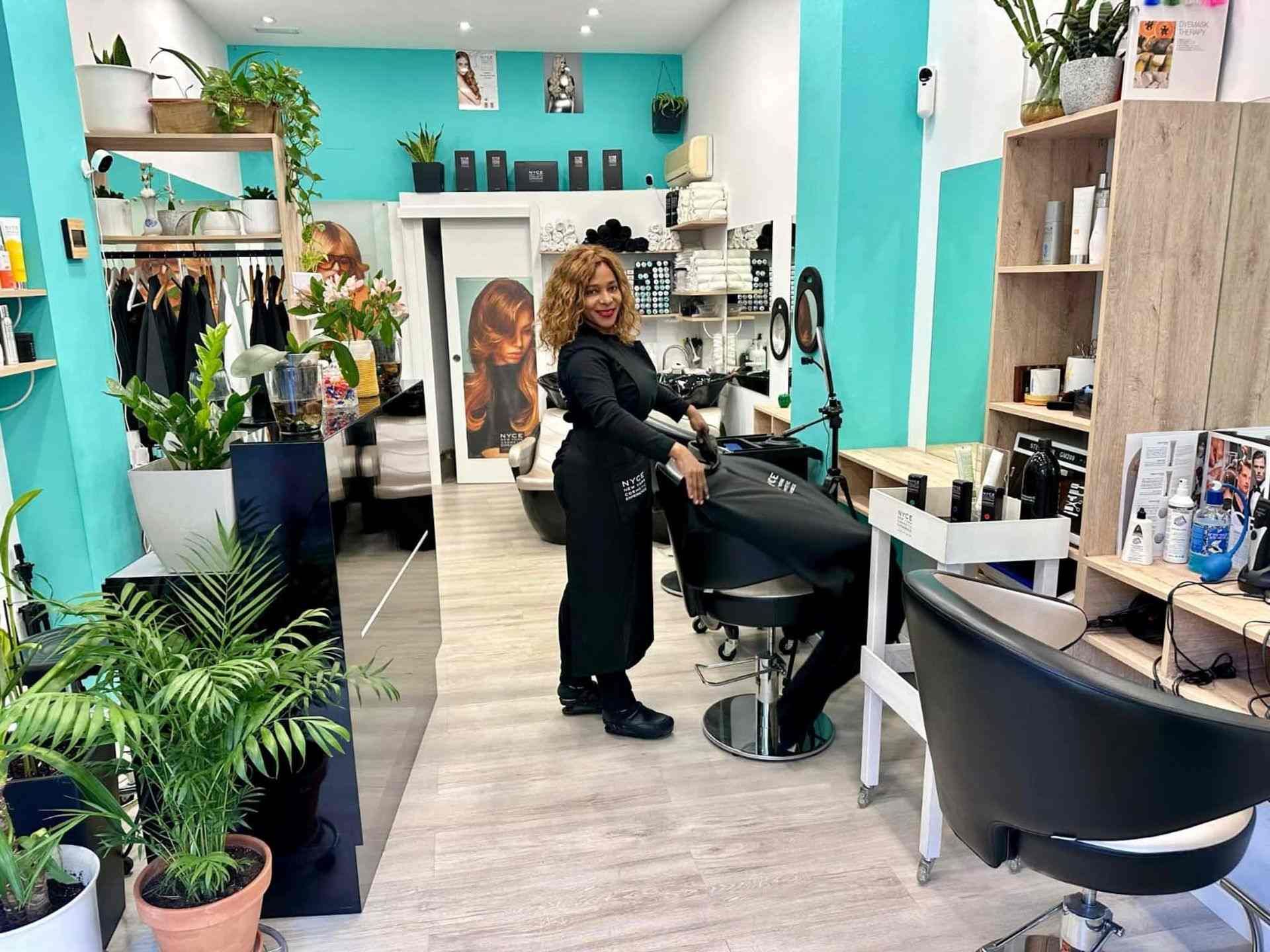 Peluquera en un salón junto a una clienta sentada. El espacio está bien iluminado con plantas y paredes turquesas.