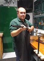 Hombre con camisa verde sosteniendo una escopeta en un taller junto a un armario y un banco de trabajo verdes.