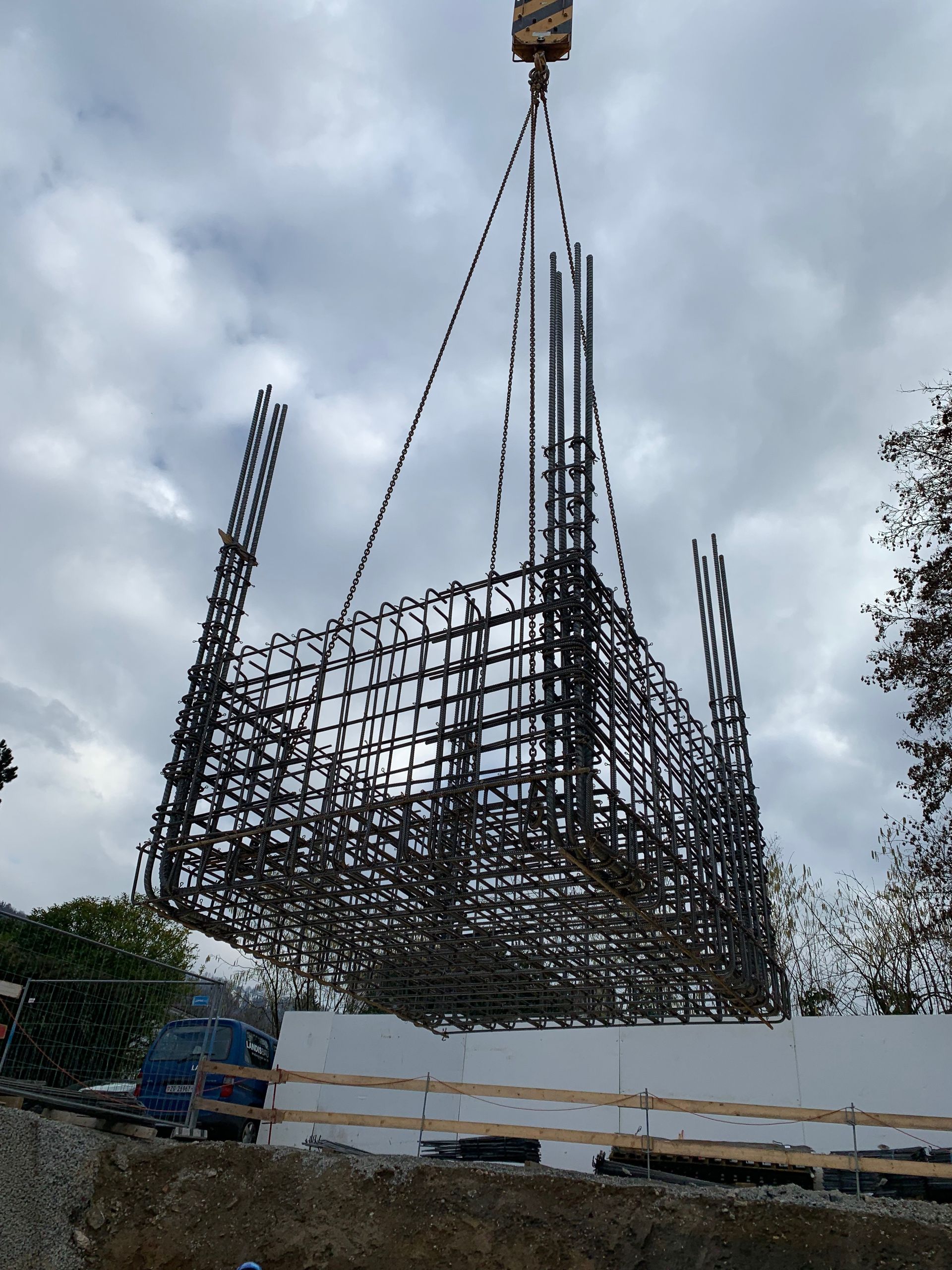 Ein Kran hebt einen großen, rechteckigen Bewehrungskorb aus Stahl auf einer Baustelle an, der Himmel ist bewölkt.