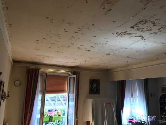 Salon avec plafond orné de taches, fenêtres ouvertes avec rideaux et meubles encombrés près d'un balcon donnant sur la vue