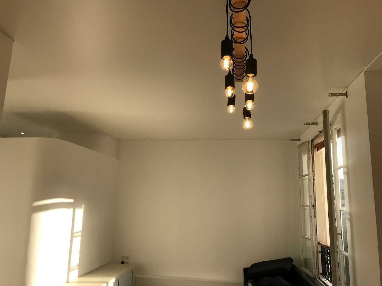 Pièce vide aux murs blancs, parquet, luminaires suspendus et chaise noire près de la fenêtre.