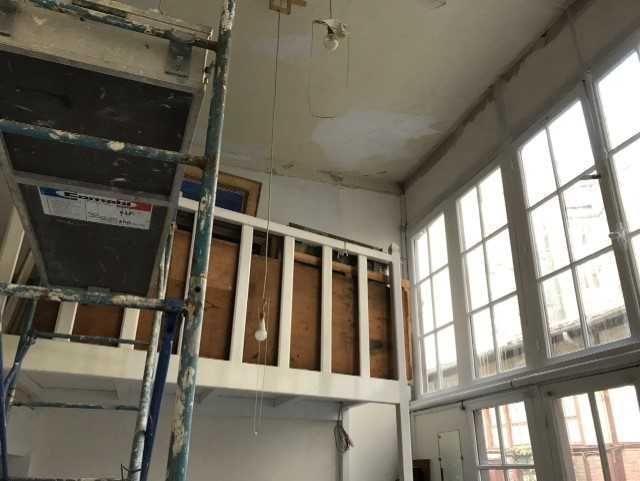Intérieur en rénovation avec échafaudages, ossature murale apparente et hautes fenêtres.