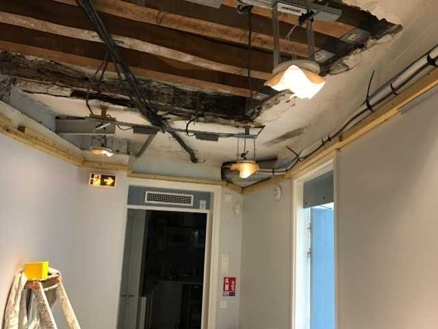 Plafond endommagé avec poutres et câbles apparents au-dessus d'une porte dans un couloir, avec une échelle à proximité.