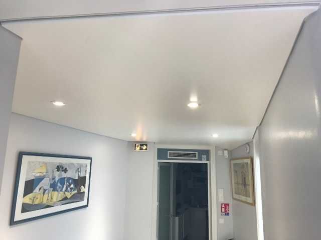 Plafond blanc dans le couloir avec deux spots encastrés, tableaux encadrés et une porte ouverte au fond.