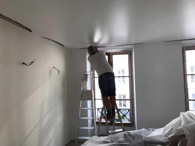 Une personne sur une échelle peint un plafond blanc dans une pièce avec des fenêtres.