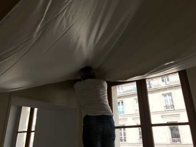 Une personne se penche pour ajuster un grand auvent ou un tissu blanc près d'une fenêtre.