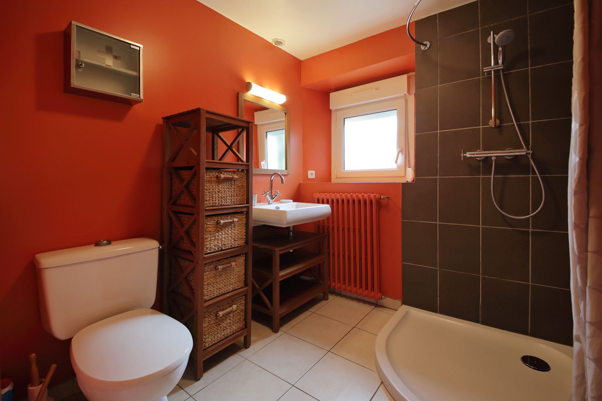 Salle de bain orange avec toilettes, lavabo, douche, fenêtre et meuble de rangement en osier