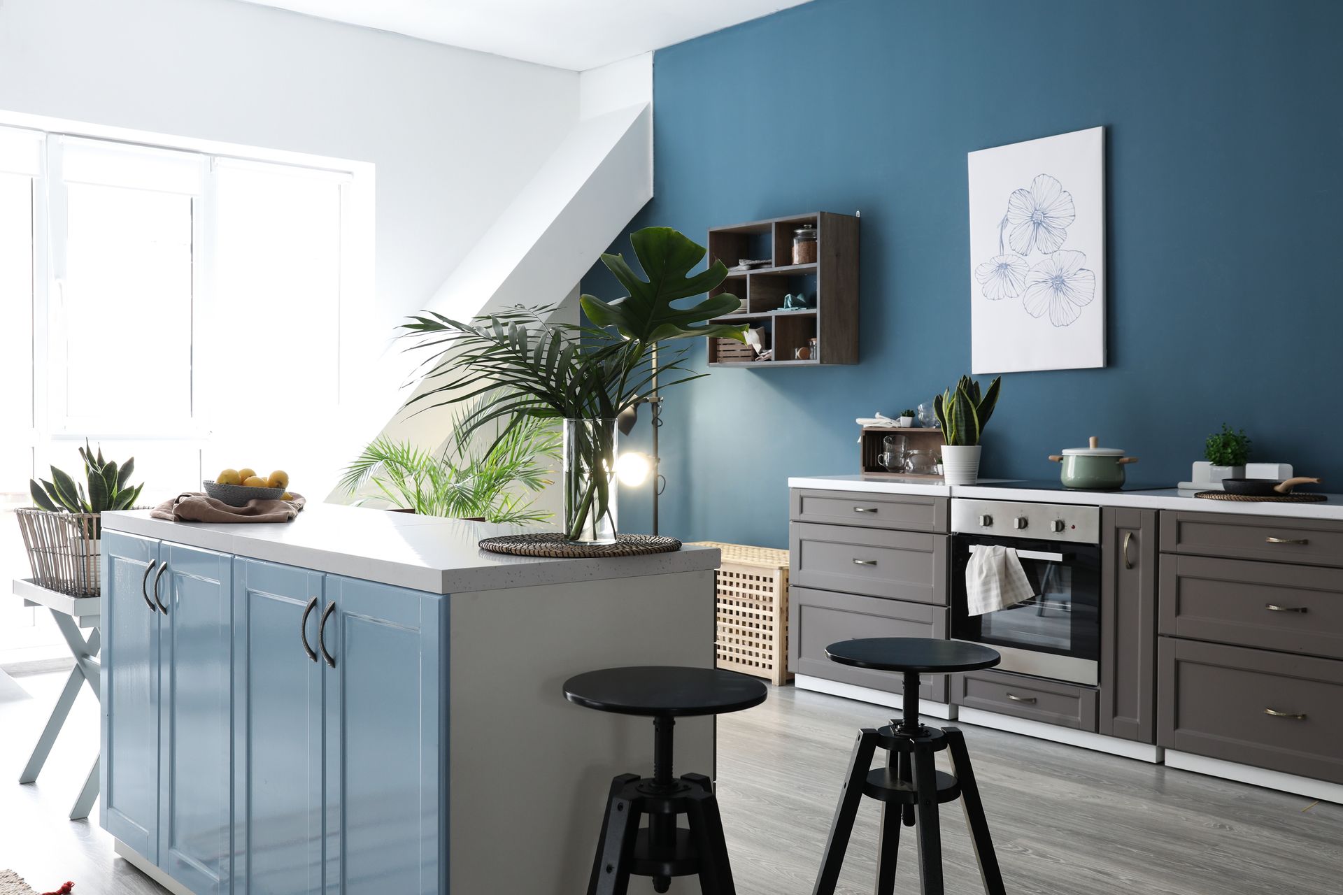 Cuisine moderne et lumineuse avec îlot bleu, placards gris, tabourets noirs et plantes près d'une fenêtre ensoleillée
