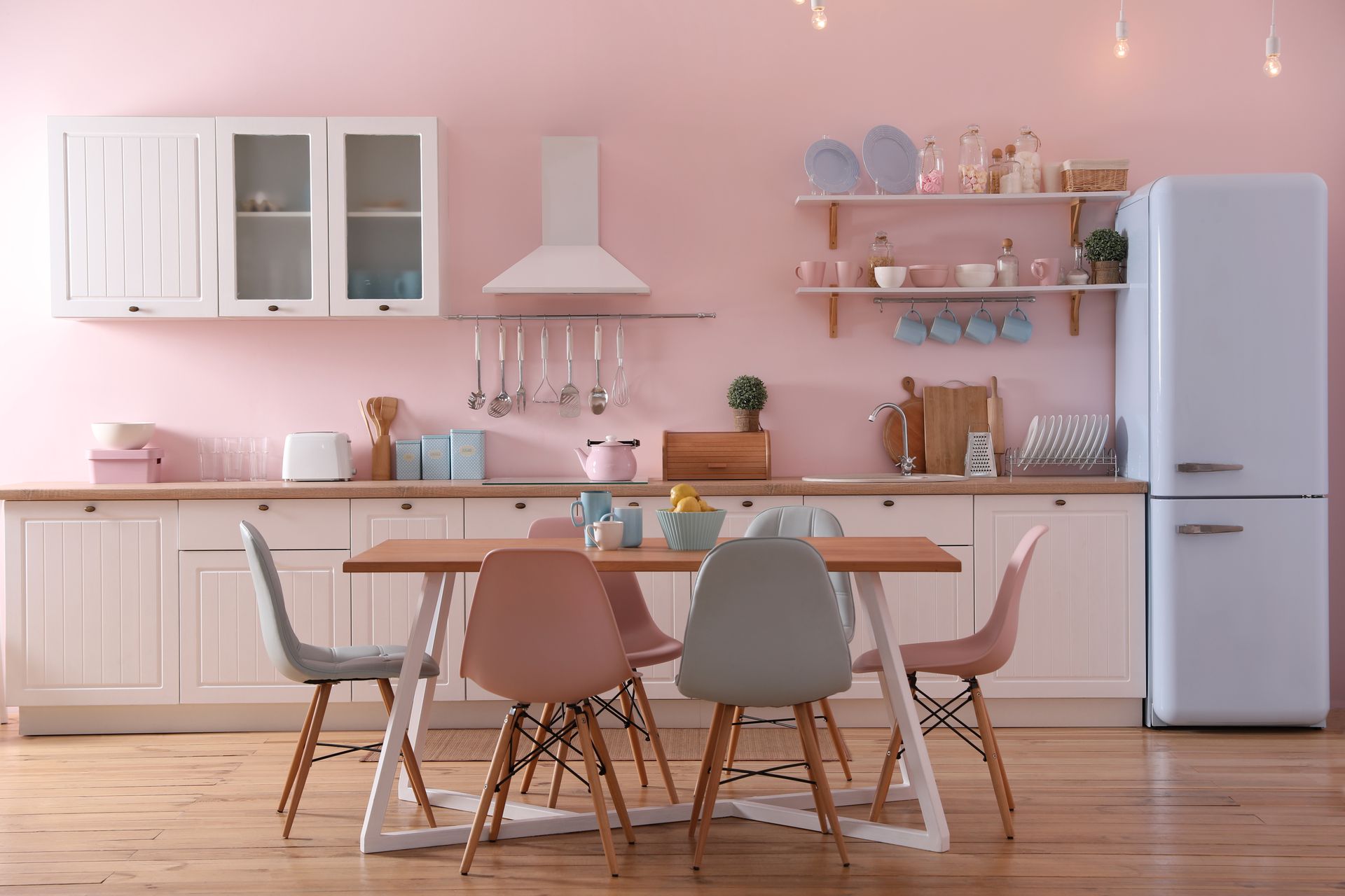 Cuisine rose pastel avec table en bois, chaises dépareillées, placards blancs et réfrigérateur rétro
