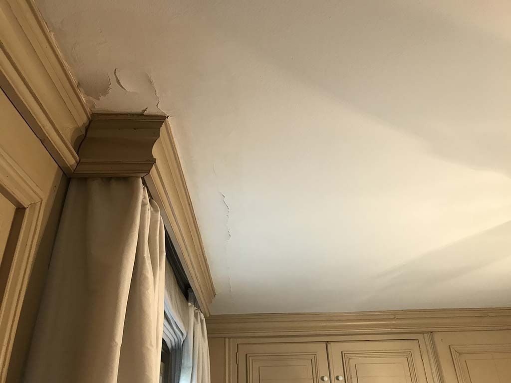 Coin d'une pièce beige avec rideaux, moulures de placard et coin de plafond blanc présentant une petite fissure.