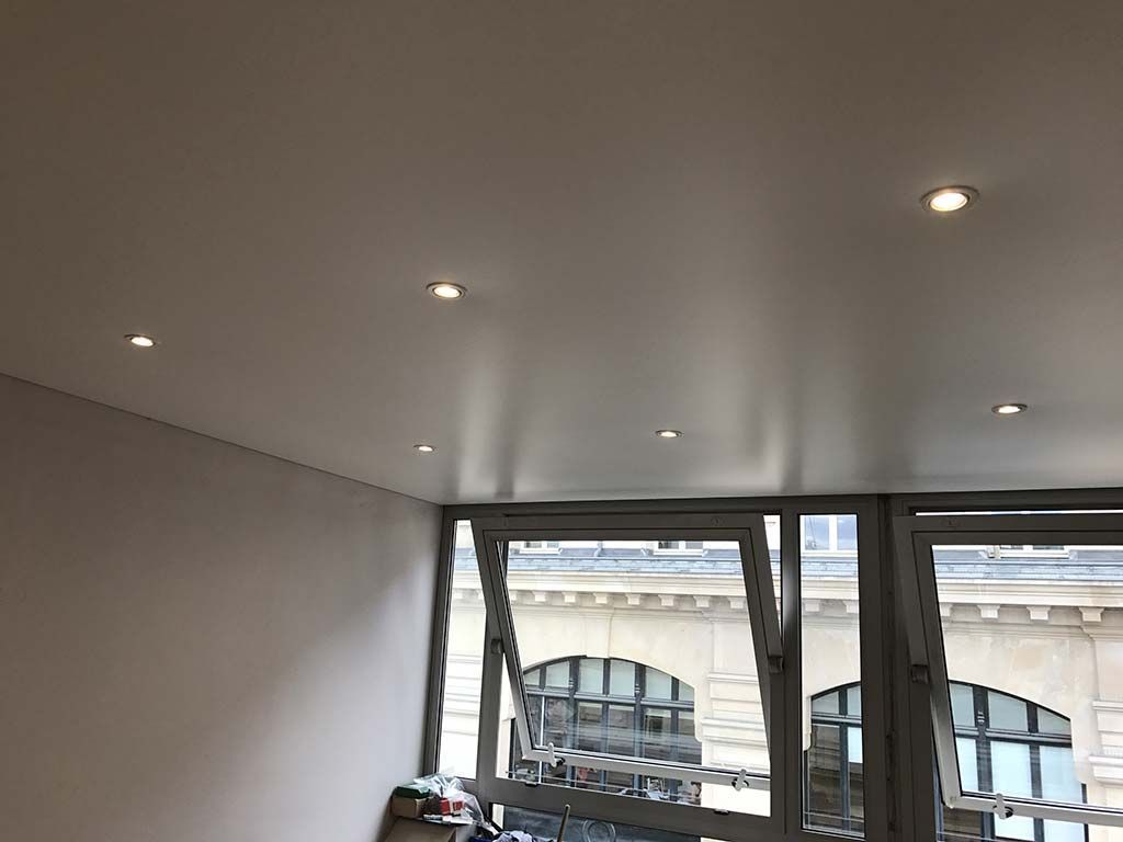 Plafond blanc avec spots encastrés au-dessus d'une fenêtre ouverte et vue sur la ville depuis les toits.