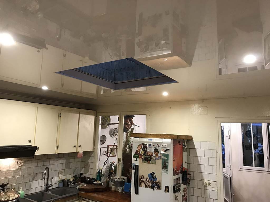 Cuisine encombrée avec des placards blancs, un réfrigérateur recouvert de photos et un panneau de plafond ouvert au-dessus.