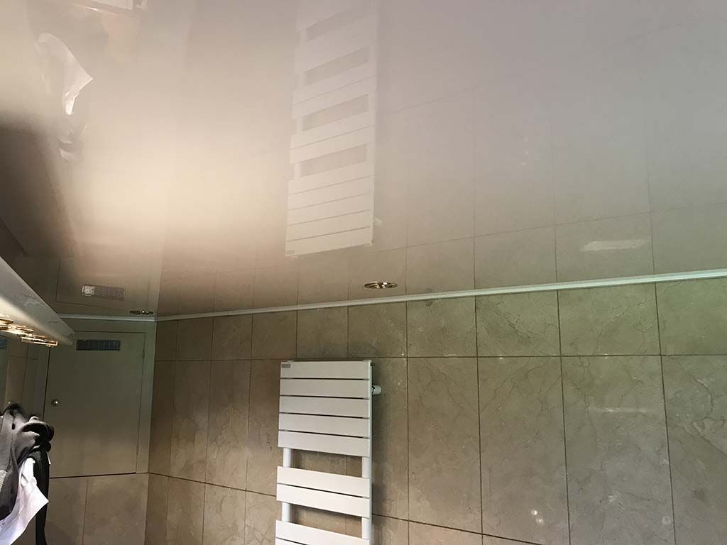 Salle de bains brumeuse avec murs carrelés beiges, un miroir et une grille d'aération murale près du plafond.