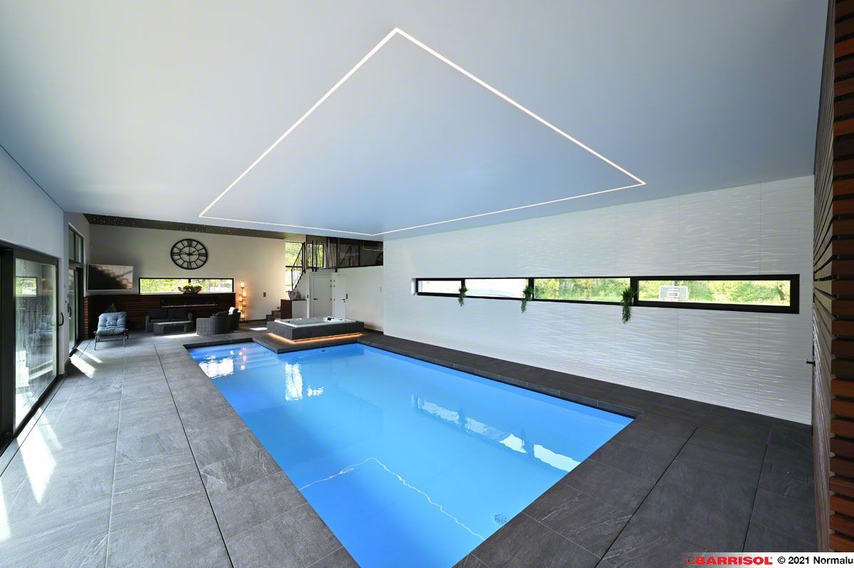 Piscine intérieure moderne avec eau bleue, terrasse en carrelage gris et luminaires triangulaires blancs au plafond.