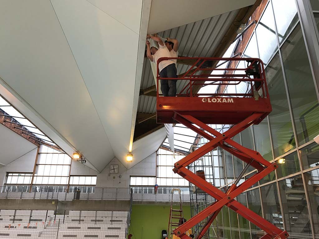 Une personne sur une nacelle élévatrice rouge travaille près du plafond à l'intérieur d'un grand bâtiment industriel.