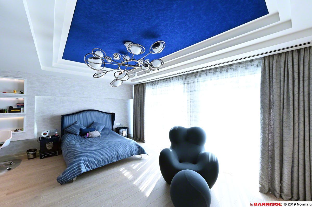 Chambre moderne avec linge de lit bleu, murs blancs, lustre, grande fenêtre et fauteuil noir près du lit.