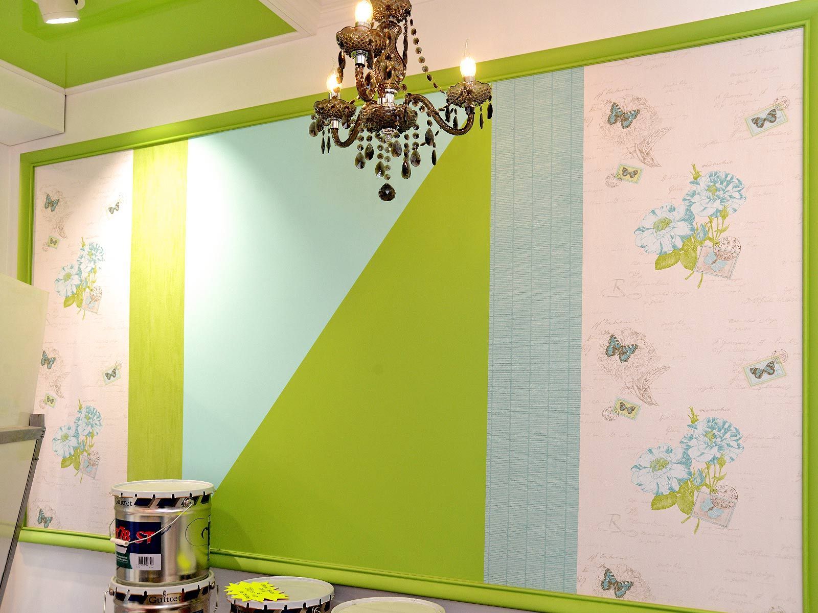 Mur d'une chambre tapissé de papier peint vert vif et pastel, avec un lustre et un pot de peinture posé au sol.
