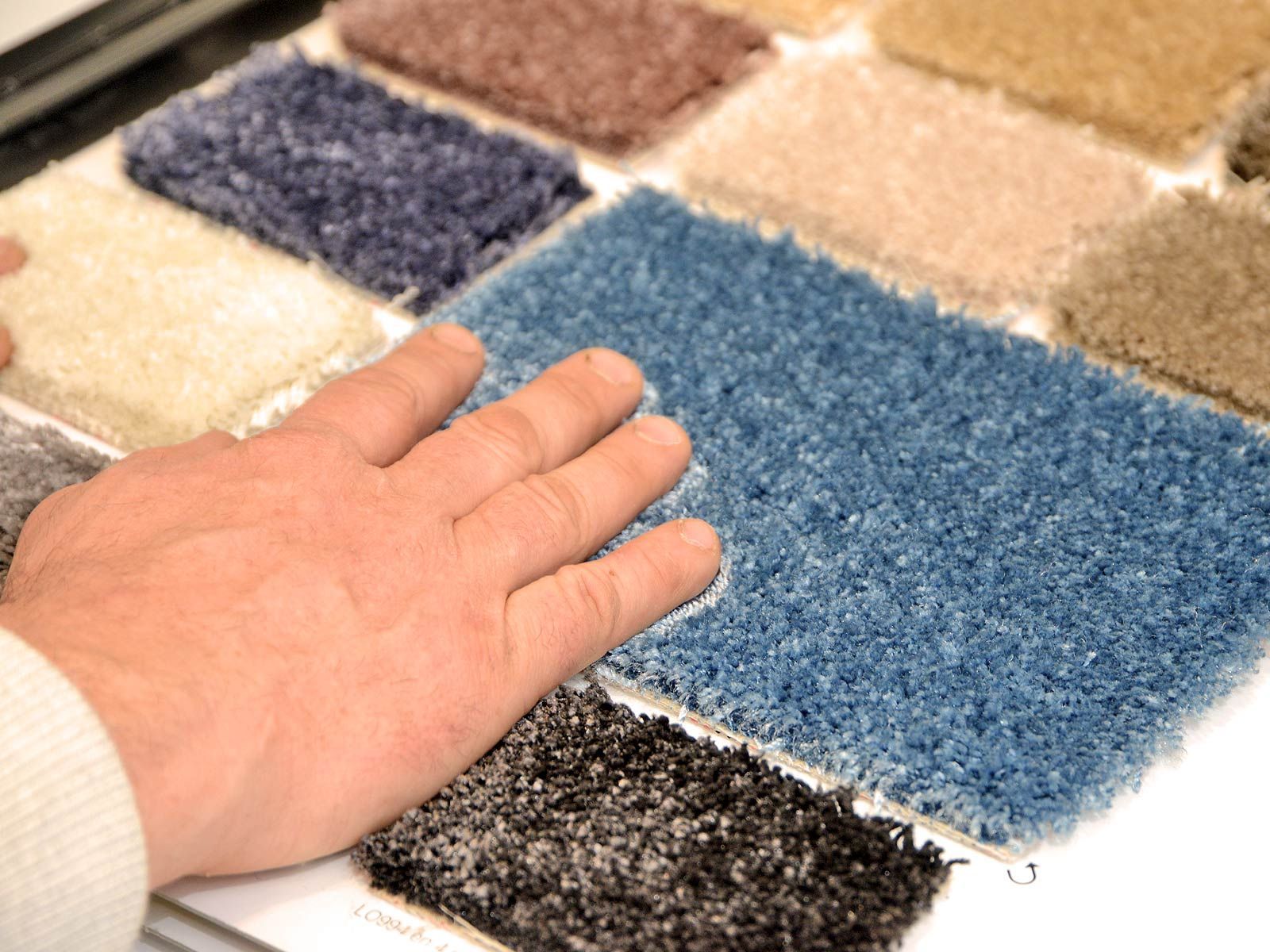 Main touchant un carreau de moquette bleue à poils longs parmi des échantillons beiges, noirs et marron clair