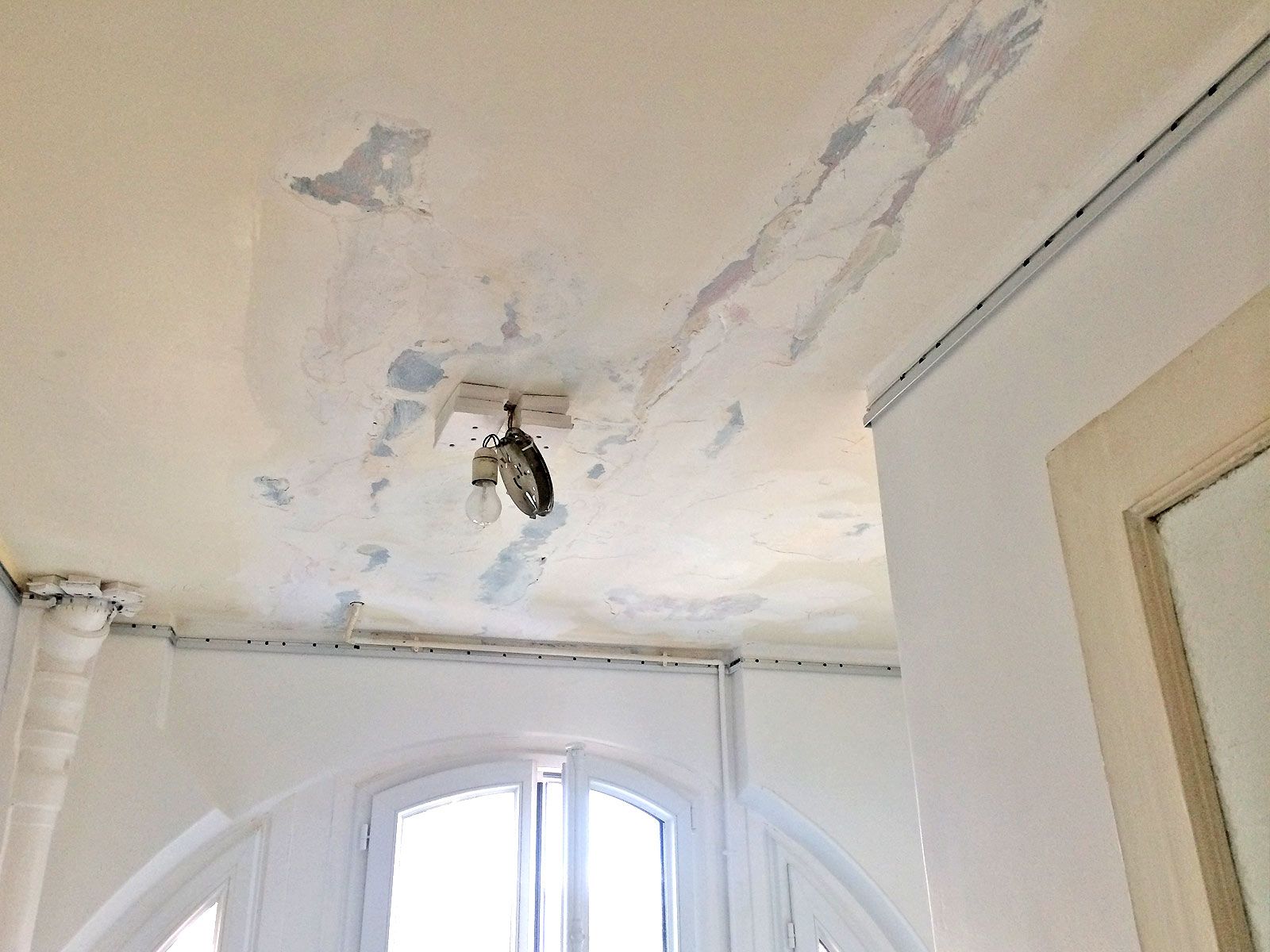 Peinture du plafond qui s'écaille au-dessus d'une fenêtre, avec un luminaire suspendu et des dégâts d'eau visibles.