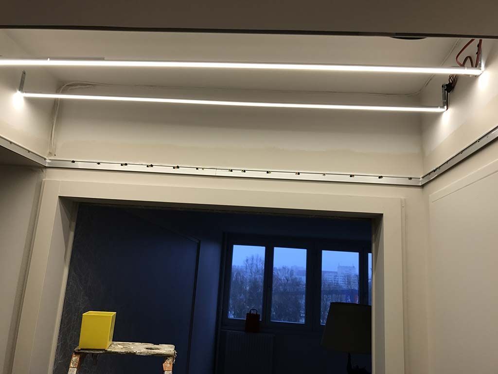 Pièce blanche en rénovation avec des luminaires apparents au plafond, une fenêtre et un seau jaune au sol.