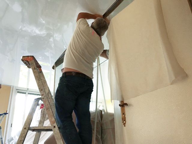Une personne, debout sur une échelle, peint un mur près du plafond dans une pièce en rénovation.