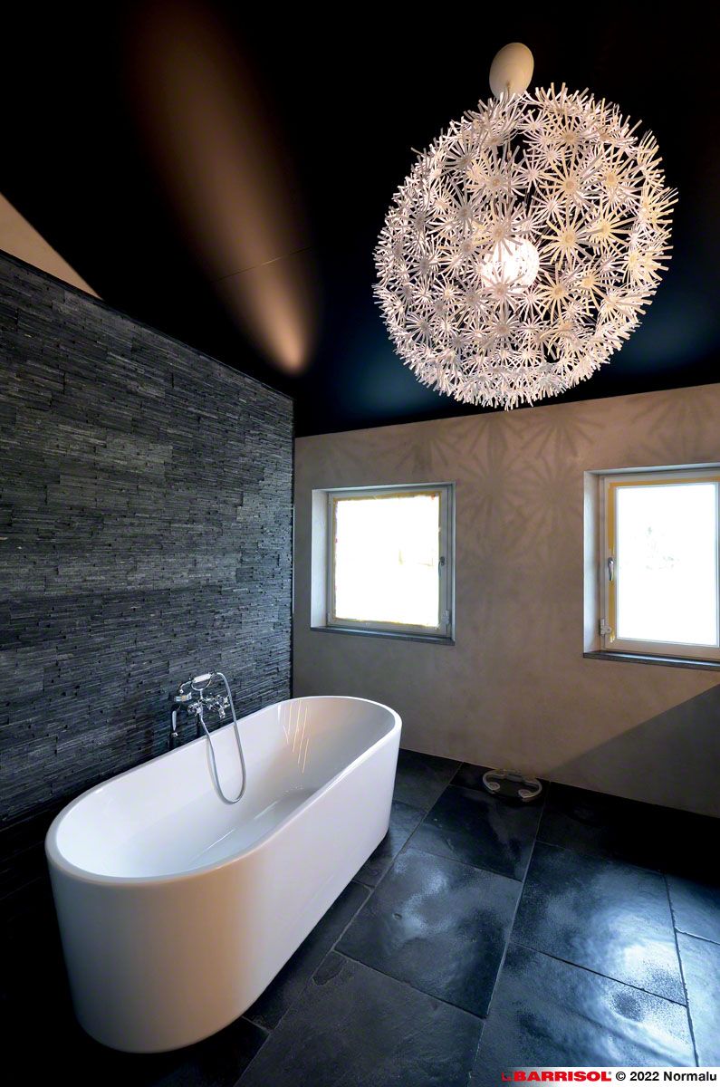 Salle de bains moderne avec baignoire îlot, sol noir, deux fenêtres et un grand luminaire suspendu.