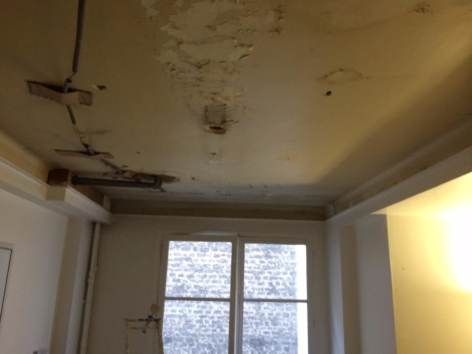 Plafond abîmé avec peinture écaillée et taches au-dessus d'une fenêtre dans une pièce peu meublée