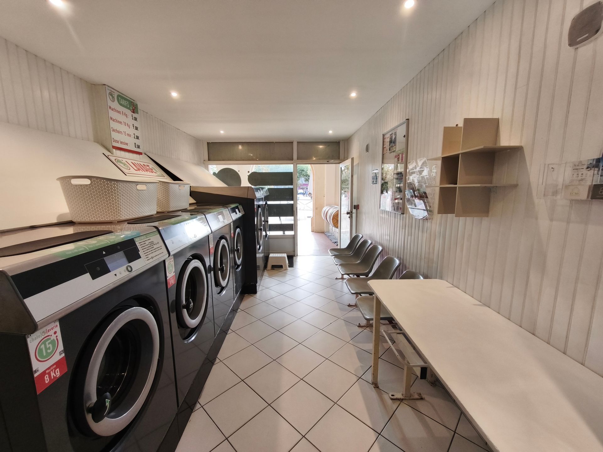 Laverie avec des lave-linge en ligne, des tables de pliage et des chaises le long d’une étroite allée carrelée.
