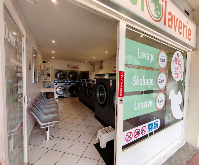 Intérieur d’une laverie avec des machines à laver, des sèche-linge et des chaises près de l’entrée.