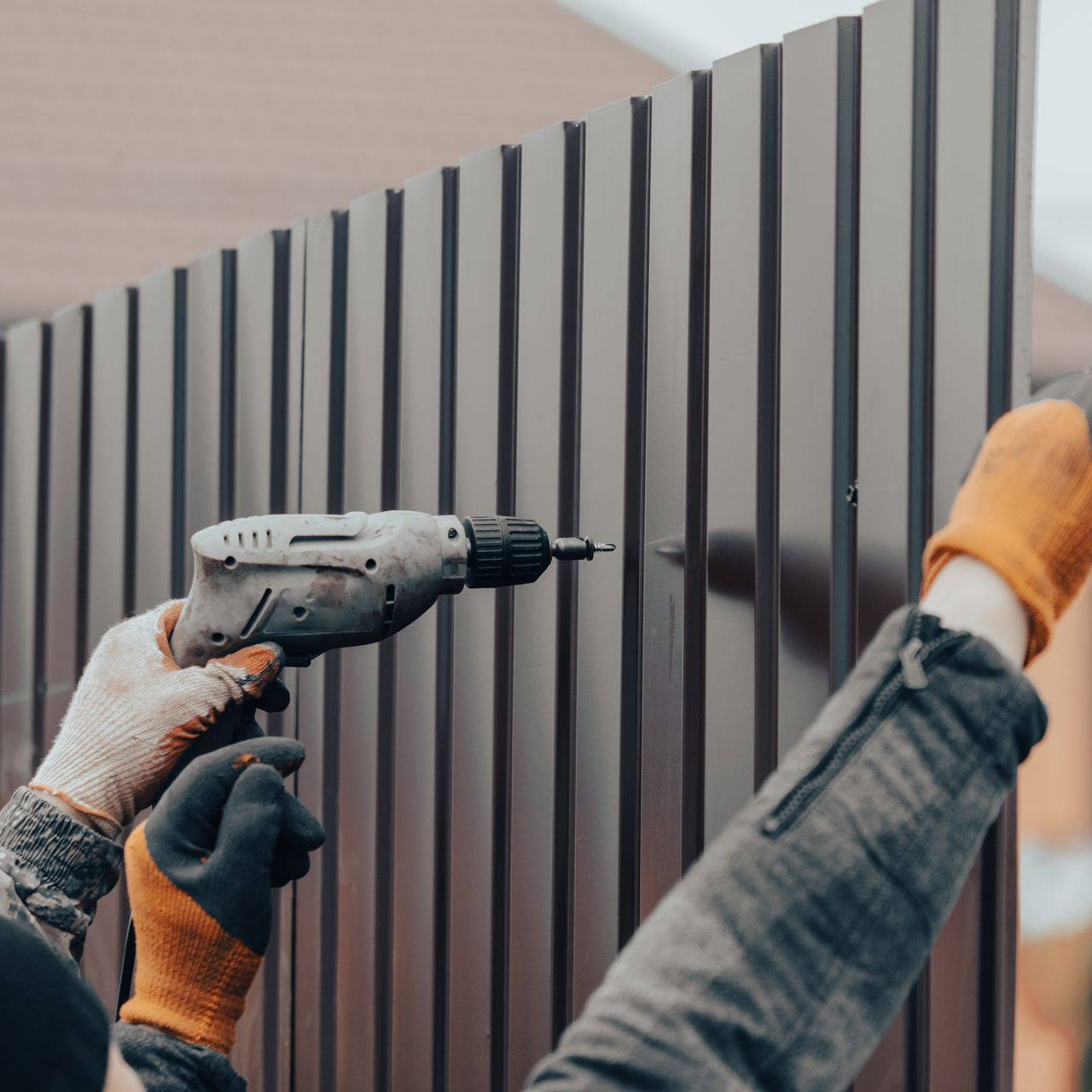Une personne portant des gants de travail utilise une perceuse électrique pour fixer des panneaux métalliques foncés sur une clôture ondulée.
