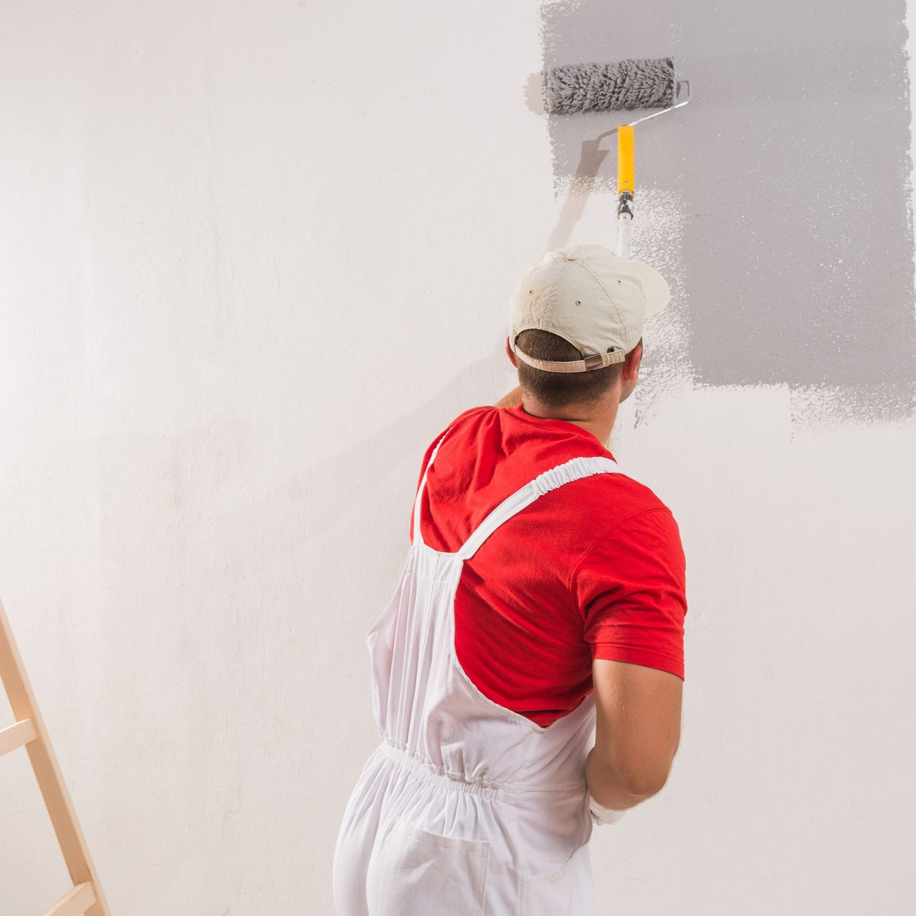 Une personne vêtue d'une salopette blanche et d'une chemise rouge utilise un rouleau à peinture pour appliquer de la peinture grise sur un mur blanc.