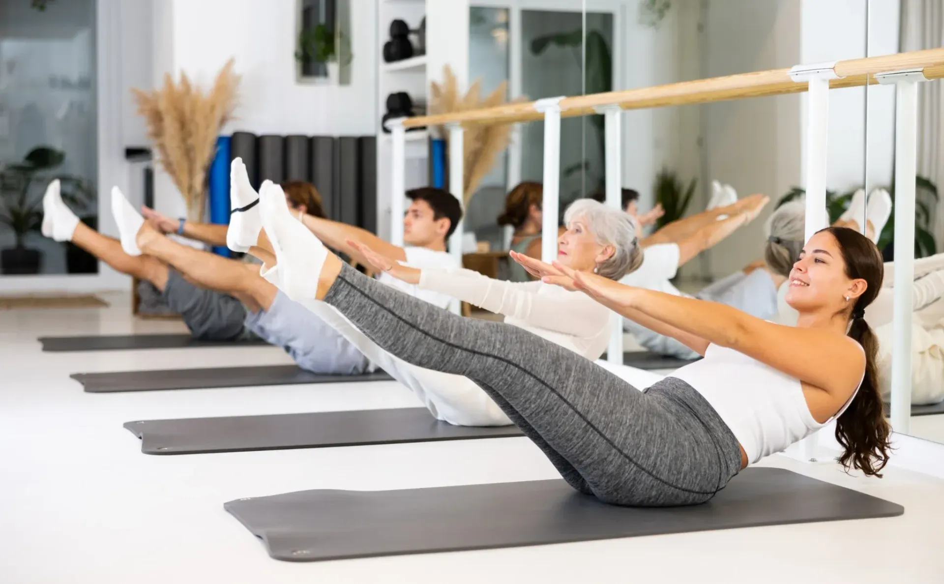 Personas haciendo ejercicios de Pilates sobre colchonetas en un estudio, extendiendo los brazos hacia adelante con las piernas levantadas.