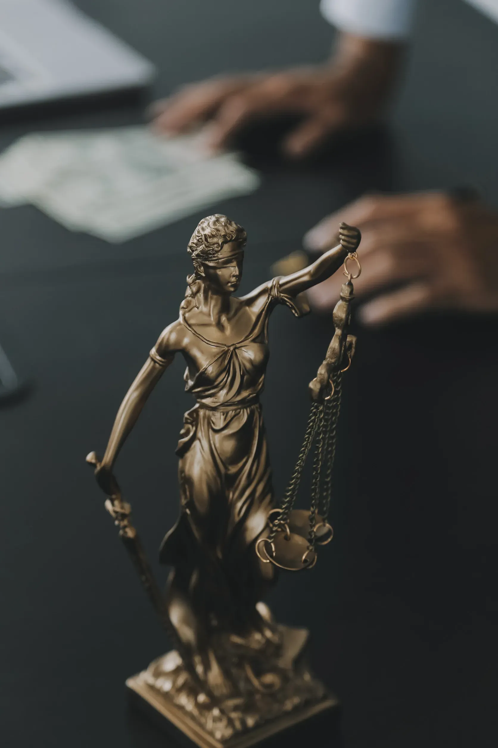 Estatua de bronce de la Dama de la Justicia en primer plano, manos borrosas y documentos sobre un escritorio detrás de ella.