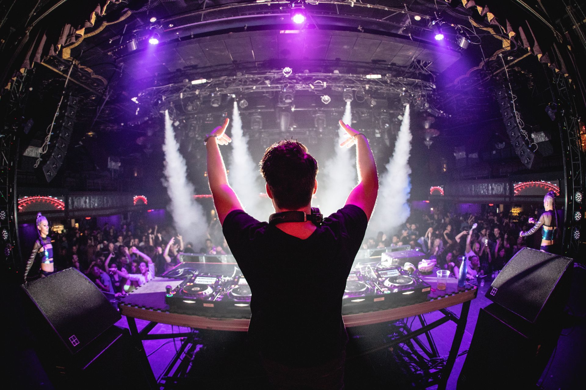 Le DJ sur scène lève les bras en direction d'une foule en liesse sous des lumières de concert et des effets pyrotechniques.