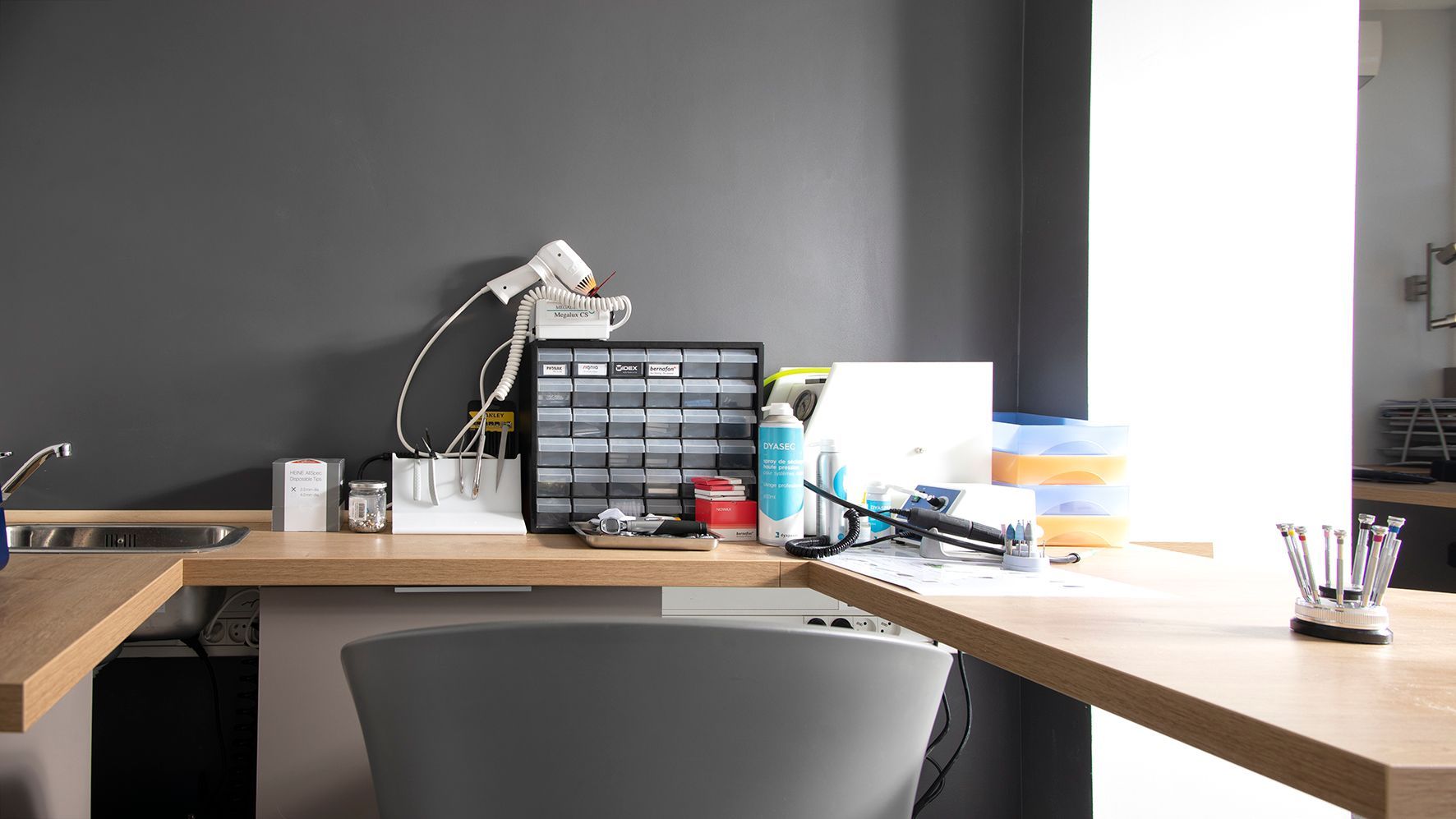 Espace de travail moderne avec lampe de bureau, fournitures rangées et chaise, adossé à un mur sombre.